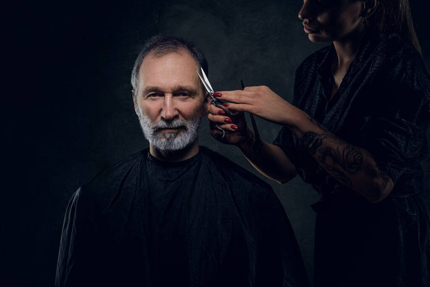 Retrato de mujer peluquera cortando pelos de su cliente mayor sobre fondo oscuro. - Foto, Imagen