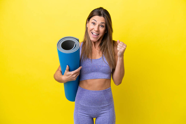 Sport woman going to yoga classes while holding a mat isolated non yellow background celebrating a victory in winner position - Photo, Image