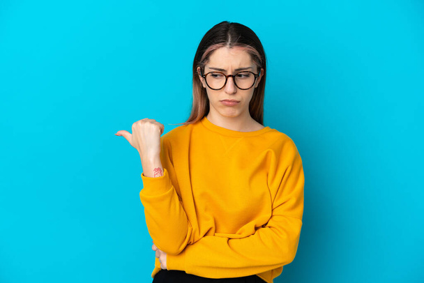 Young caucasian woman isolated on blue background unhappy and pointing to the side - Photo, Image