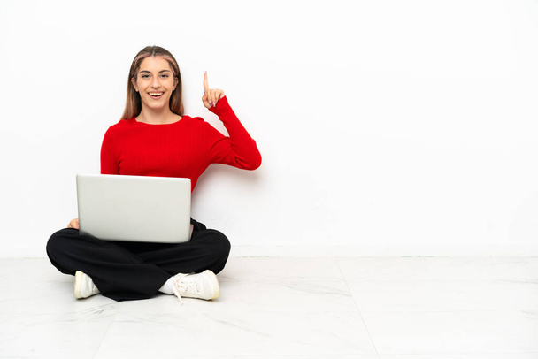 Young caucasian woman with a laptop sitting on the floor intending to realizes the solution while lifting a finger up - Photo, Image