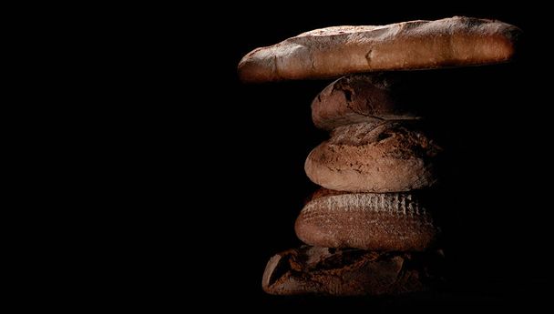 Delicious freshly baked bread loaves and baguettes stacked on top of each other on a black background - Fotografie, Obrázek