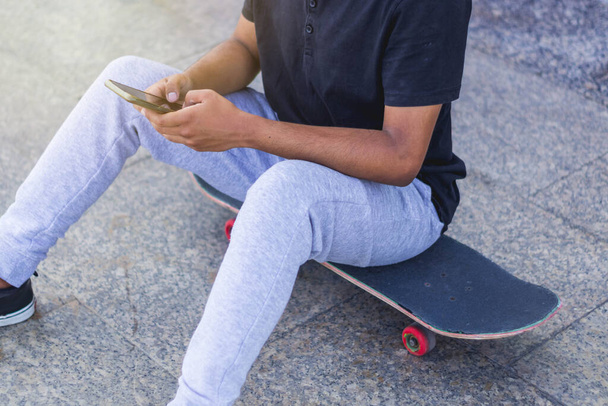 Σκούρο δέρμα τύπος κάθεται σε ένα skateboard και χρησιμοποιώντας το κινητό του τηλέφωνο. - Φωτογραφία, εικόνα