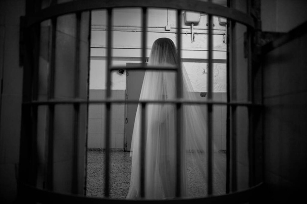A greyscale photo of a bride behind iron bars in a dark room - Foto, Imagem