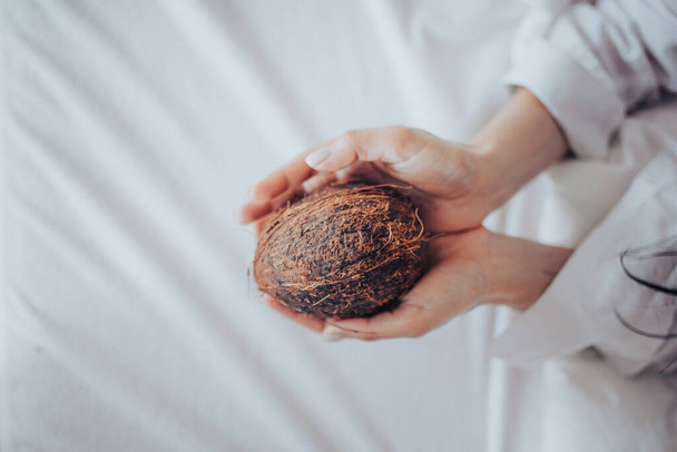 A vertical shot of a woman holding a dehusked coconut on a white fabric surface - Foto, Imagen