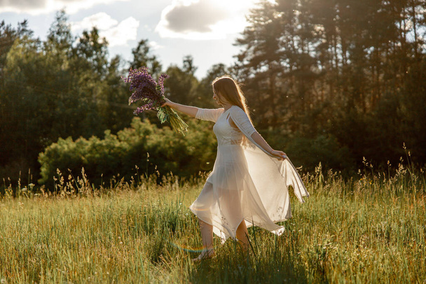 Young beautiful woman, wearing white dress, holding flowers and dancing on the meadow. Girl joying nature and freedom. Natural beauty. Dance, movement. Mental health, stress free, dreaming. Sunset - Foto, afbeelding