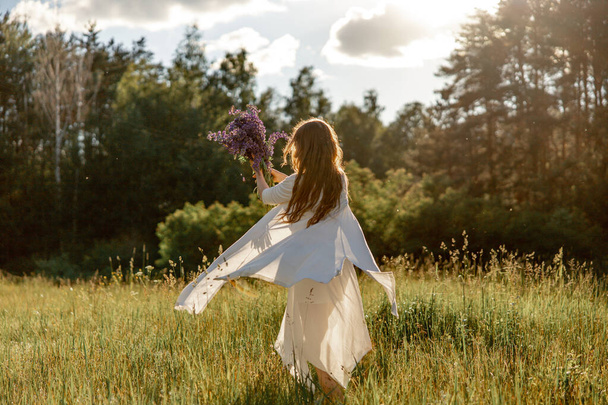 Jovem mulher bonita, vestindo vestido branco, segurando flores e dançando no prado. Menina desfrutando da natureza e da liberdade. Beleza natural. Dança, movimento. Saúde mental, sem stress, a sonhar. Pôr do sol - Foto, Imagem