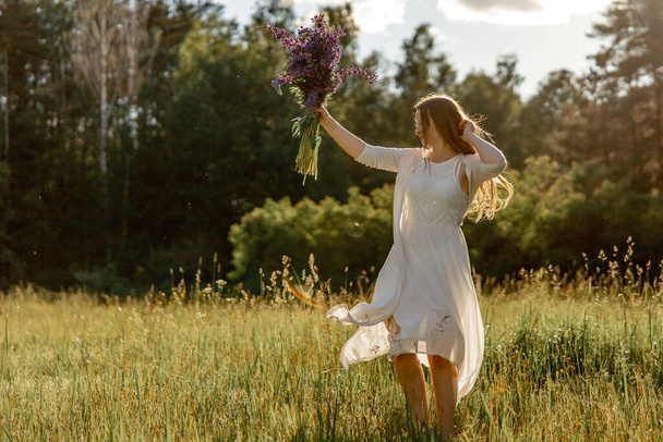 Joven mujer hermosa, vistiendo vestido blanco, sosteniendo flores y bailando en el prado. Chica disfrutando de la naturaleza y la libertad. Belleza natural. Baila, mueve. Salud mental, sin estrés, soñando. Puesta de sol - Foto, imagen