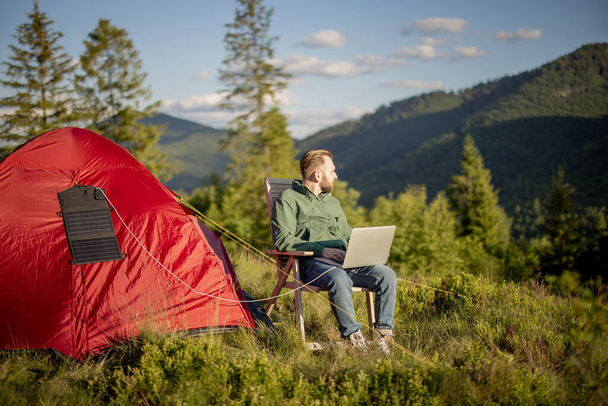 Homme travaille sur ordinateur portable tout en voyageant avec tente dans les montagnes. Ordinateur de charge avec panneau solaire portable suspendu à la tente. Concept de télétravail au camping et d'énergies renouvelables - Photo, image