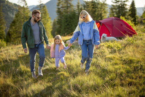 Joven familia caucásica con niña caminan juntos en el prado verde mientras viajan con tienda de campaña en las montañas. Felices vacaciones familiares de verano en el camping - Foto, imagen