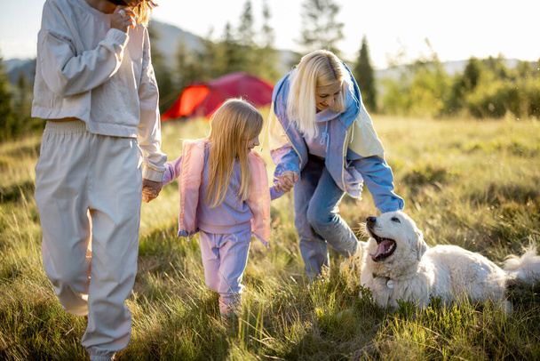 Two women walk with little girl and white dog, happily spending summer time on nature. Homosexual family with a kid travel in mountains - Photo, Image