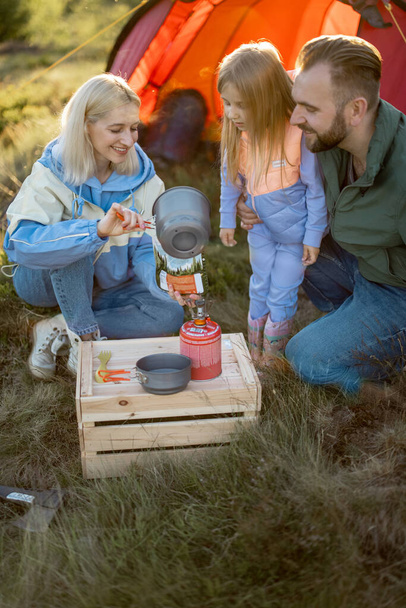 Jonge echtpaar met klein meisje koken gedroogde-voedsel voor wandelen, gieten kokend water in het pakket. Familie op de camping maakt eten voor reizigers. Gesublimeerde voeding en familie camping concept - Foto, afbeelding