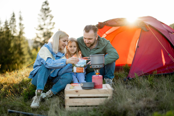 Молодая пара с маленькой девочкой устраивает пикник, готовит сушеную пищу для похода в упаковке в кемпинге. Концепция сублимационного питания для путешествий и семейного сближения в путешествиях - Фото, изображение