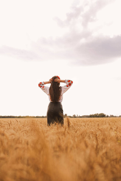 Ein junges hübsches Mädchen steht auf dem Feld und trägt ein besticktes ukrainisches Nationalhemd mit rotem Kranz. Konzept der Ukraine Unabhängigkeit, Freiheit und Beendigung des Krieges. Frieden in Worten. Sonnenuntergang gelbes Sommerlicht - Foto, Bild