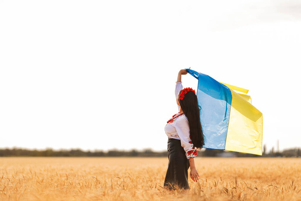 Joven chica bonita sostiene la bandera y la espera de la victoria en el campo con la camisa bordada nacional ucraniana con corona roja. Concepto de Ucrania Independencia, libertad y detener la guerra. Paz en la palabra - Foto, imagen