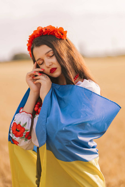 Mladá smutná dívka zahalená do vlajky, čekající na vítězství v poli, oblečená do ukrajinské národní vyšívané košile s červeným věncem. Koncept Ukrajiny Nezávislost, svoboda a zastavení války. Mír ve slově - Fotografie, Obrázek
