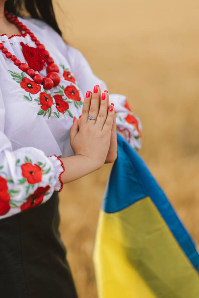 Young girl prays in the wheat field wearing Ukrainian national embroidered shirt with flower red wreath and holds flag at sunset yellow light. Concept Independence, freedom of Ukraine and stop the war - Photo, Image