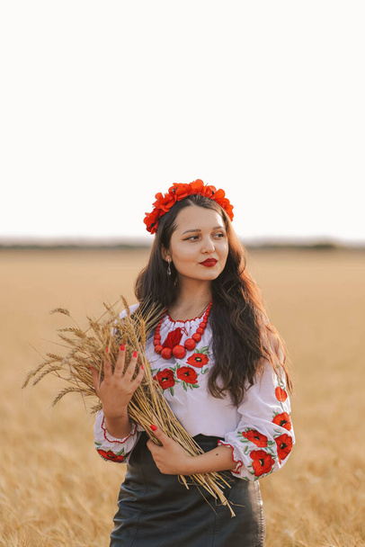 Young girl in the field wearing Ukrainian national embroidered shirt with red wreath and holds wheat bouquet. Concept of Ukraine freedom, independence and stop the war. No hunger in the world. Peace - Zdjęcie, obraz
