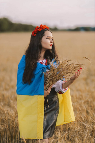 Young sad girl shrouded in flag waiting for victory in the field wearing Ukrainian national embroidered shirt with red wreath and holds wheat bouquet. Concept of Ukraine freedom and stop the war - Photo, Image