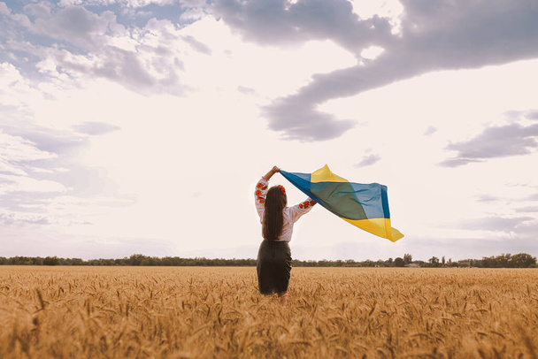 Молодая красивая девушка держит флаг и ждет победы на поле в украинской национальной вышитой рубашке с красным венком. Концепция независимости Украины, свободы и прекращения войны. Мир на словах - Фото, изображение