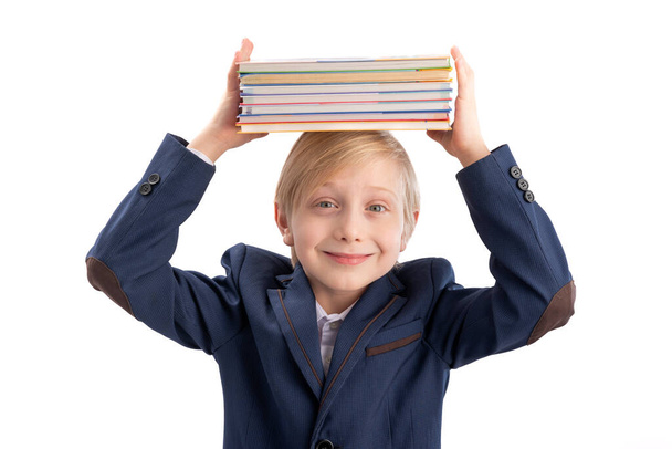 Gülümseyen ilkokul öğrencisinin portresi. Okul üniformalı öğrenci kafasında bir yığın kitap tutuyor. Beyaz arkaplanda izole - Fotoğraf, Görsel