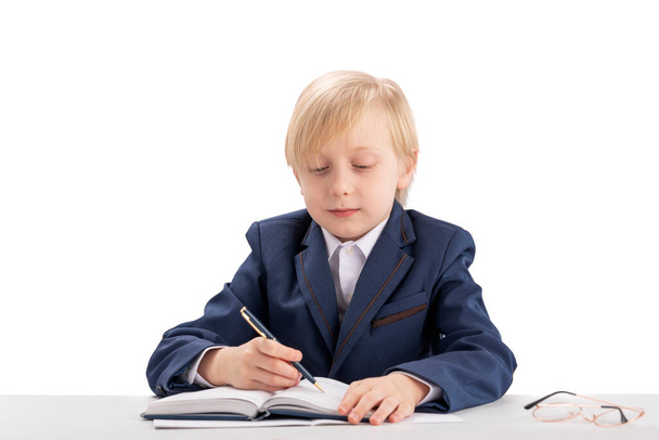 A szorgalmas diák másolóban ír. A gyerek, mint a főnök, asztalhoz ül és jegyzetfüzetbe ír. Szőke fiú zakóban és ingben. - Fotó, kép