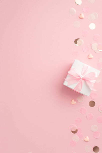 Genderpartijconcept. Bovenaanzicht verticale foto van witte geschenkdoos met lint strik harten en glanzende confetti op geïsoleerde pastel roze achtergrond - Foto, afbeelding