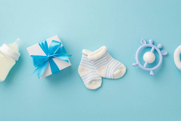 Het concept van een baby. Bovenaanzicht foto van giftbox bijtring melk fles en kleine sokken op geïsoleerde pastel blauwe achtergrond - Foto, afbeelding
