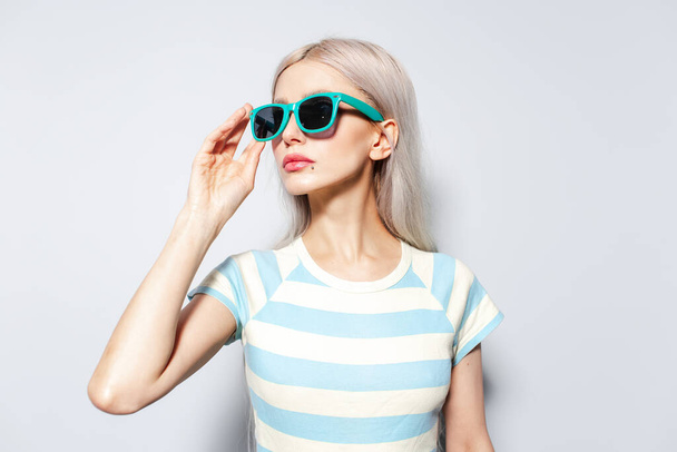 Modieus blond meisje in gestreept shirt met een zonnebril op een witte achtergrond. - Foto, afbeelding