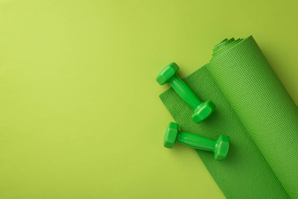 Концепция аксессуаров. Фото зеленого спортивного коврика и гантели на изолированном зеленом фоне с копирайтом - Фото, изображение
