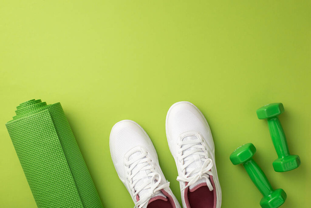 Actief woonconcept. Bovenaanzicht foto van witte sneakers groene sportmat en halters op geïsoleerde groene achtergrond met copyspace - Foto, afbeelding