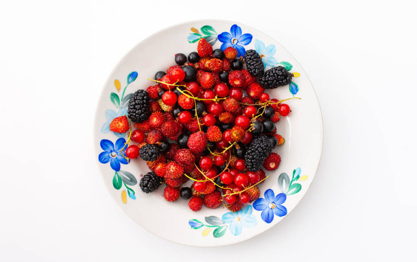 Fresh berries on a plate on a white background, red and black currants and black mulberries, wild strawberries and cherries, top view - Photo, image
