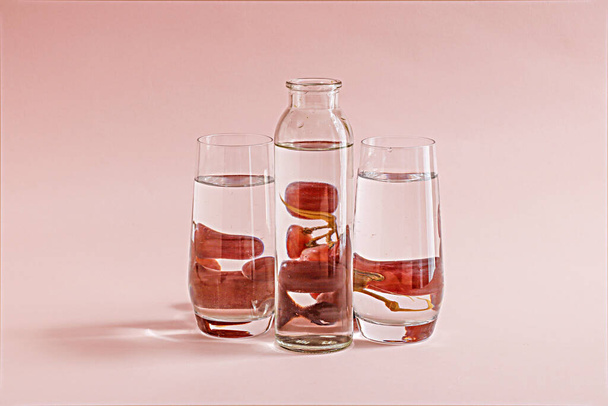 A transparent bottle of water and two glasses with grapes behind them on a pink background - Фото, изображение