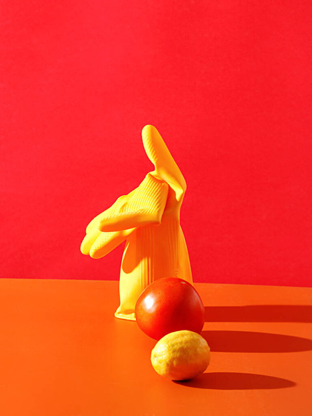 A closeup shot of fresh ripe Lemon, tomato and single heavy duty yellow rubber glove on an orange table against red wall - Фото, изображение