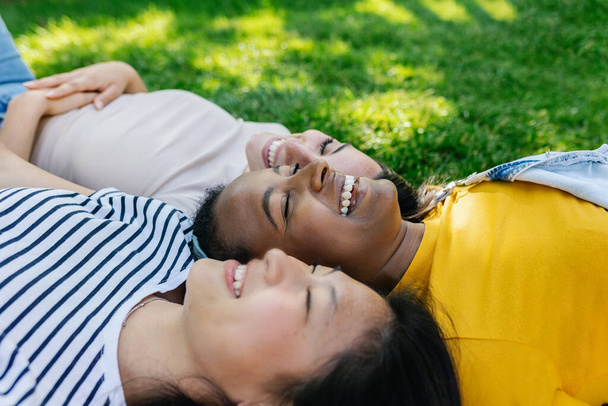Drei multiethnische junge Freundinnen, die gemeinsam Spaß im Freien haben - Weibliches Freundschaftskonzept mit einer kleinen Gruppe unterschiedlicher Teenager-Mädchen, die im öffentlichen Park auf Gras liegen und lachen - Foto, Bild