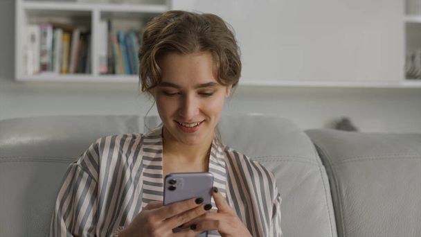 Счастливая девушка пишет смс на портрете мобильного телефона. Пользователь приложения, использующий смартфон, отдыхает на диване. Ошеломленный улыбчивый студент в гостиной. Радостная красавица наслаждается утренним серфингом в Интернете - Фото, изображение