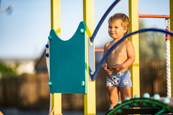 A small naked cheerful kid in bright blue shorts plays on a children's playground while walking outside on a warm sunny day, under the summer golden evening sun - Foto, Imagen