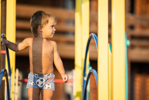 A small naked cheerful kid in bright blue shorts plays on a children's playground while walking outside on a warm sunny day, under the summer golden evening sun - Zdjęcie, obraz