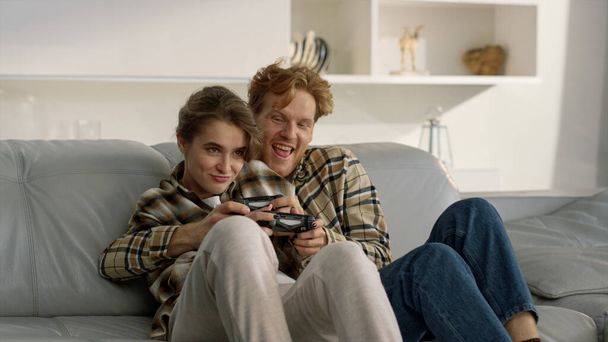 Emotional friends play gamepad video game at home. Joyful family time together. Happy woman excited wife cheating winning online tournament. Playful girl biting redhead man hand celebrating victory. - Foto, Imagem
