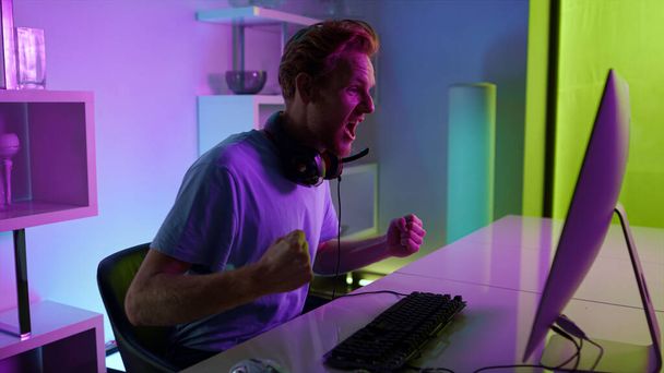 Cyber game winner enjoying success in neon room. Excited student rest on weekend night. Happy ginger man shouting making yes gesture at home. Professional gamer celebrating triumph online victory. - Foto, Imagen