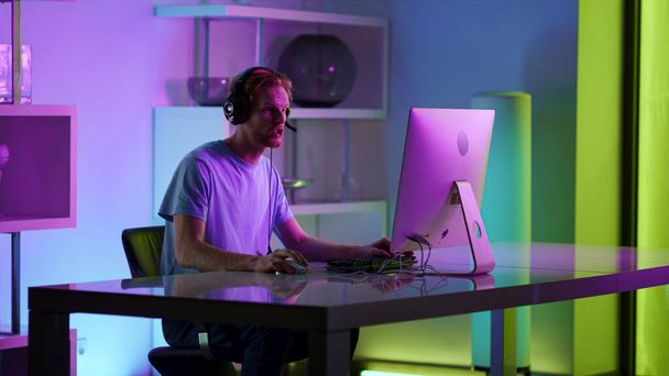 Cyber játékos beszél mikrofon otthon. Esport streamer kommentálja a neon fényeket. Jóképű fókuszált férfi játékos élvezni számítógépes játék este headset. Ginger srác az online lövöldözős versenyen. - Fotó, kép