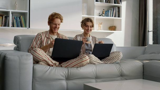 Happy couple using laptops resting on weekend. Young people work in home office. Two smiling freelancers talk drinking coffee enjoying morning together on couch. Ginger man watch checking social media - Photo, Image