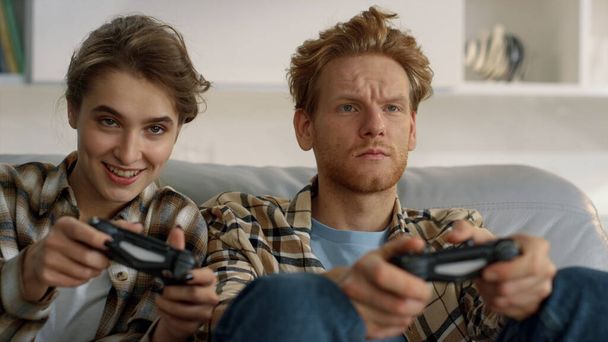 Joyful wife holding gamepad fighting confused redhead husband at home closeup. Excited woman enjoying virtual competition with frustrated ginger man. Friends using joysticks play video game on weekend - Photo, Image