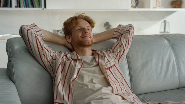 Smiling guy lean couch at cozy home closeup. Happy freelancer resting on couch enjoying siesta. Young ginger man having break taking nap in pajamas. Satisfied manager student relax chilling on weekend - Foto, Imagen
