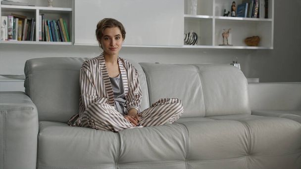 Beautiful housewife posing sofa in pajamas before breakfast. Girl relaxing alone enjoying calm morning atmosphere in lotus position at cozy home. Attractive blonde businesswoman resting on weekend. - Zdjęcie, obraz