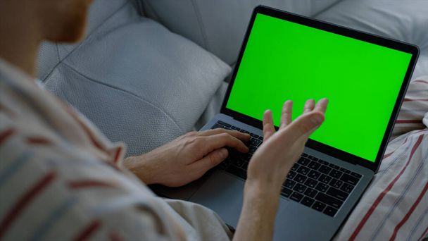 男はソファの上で休んで緑のノートパソコンを話している。リモートホームオフィスでオンライン講義をしている教師。認知されていないフリーランサーの挨拶のクライアントの友人パジャマに横たわって手を振る。モックアップ・コンピュータの概念. - 写真・画像
