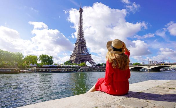 パリの有名なランドマークと旅行先、エッフェル塔を見てセーヌ川のほとりに座って赤いドレスと帽子の若い旅行者の女性. - 写真・画像