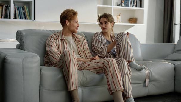 Happy couple having conversation in morning. Young family resting on weekend together. Smiling blonde girl ginger man talking discussing funny story in pajamas living room. Lovers enjoy time at home - Foto, Imagen