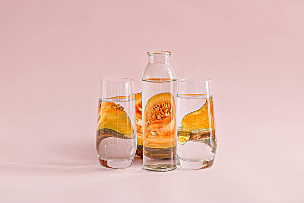 A transparent bottle of water and two glasses with a melon behind them on a pink background - Photo, image