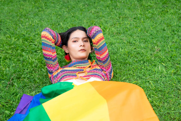 Transgender-Frau mit Regenbogenbluse, die die Regenbogenfahne unter dem Körper bedeckt, liegt auf grünen Gräsern und repräsentiert die Gleichstellungsbewegung lgbtq und den lgbtq glücklichen Stolz Monat - Foto, Bild