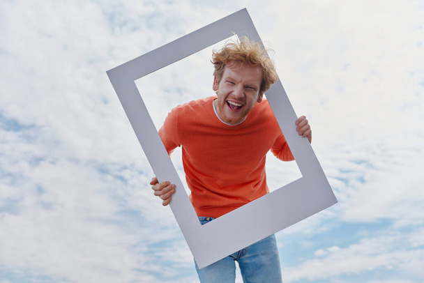 Playful young redhead man looking through a picture frame and smiling while standing outdoors - Foto, Imagen